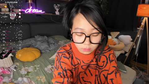 Media: Video of an Asian woman with straight black hair, wearing glasses and a patterned orange sweater, lying on a messy bed with stuffed animals and a floral blanket. Background includes a purple light and a lamp.