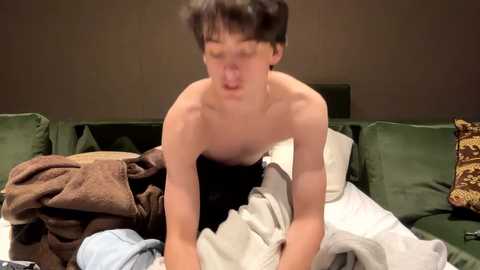 Media: A young, shirtless Asian male with short black hair, wearing black pants, lies on a bed with messy white and green blankets, making a humorous face.