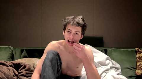 Media: Video of a shirtless young man with short dark hair, sitting on a bed with green cushions and rumpled white sheets, in a dimly lit room.