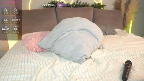 Media: Video of a cozy bedroom with a large, light blue pillow on a white, knitted blanket. A pink pillow and a brown object (possibly a remote control) are visible on the bed.