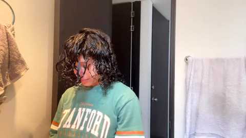 Media: Video of a person with curly black hair, wearing a teal shirt with \"Los Angeles\" printed in white, standing in a bathroom with a towel rack and door.