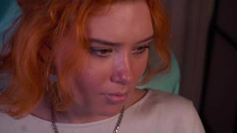 Media: Video of a young woman with fair skin and red hair, wearing a white top, looking contemplative. Background is blurred, suggesting a dimly lit room.