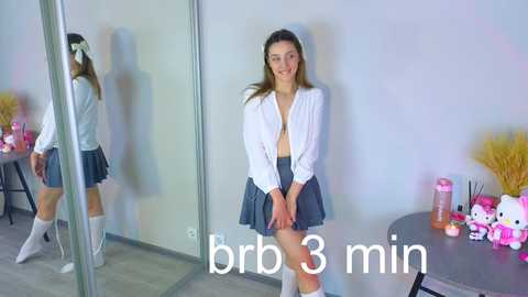 Media: Video of a smiling young woman with light skin, brown hair, and medium build, wearing a white blouse and short skirt, standing in front of a mirror.