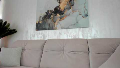 Media: Video of a modern living room with a light grey tufted sofa, a large abstract painting in muted colors, and a green plant in the corner.