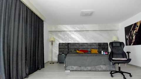 Media: Video of a modern bedroom with a grey bed, black curtains, white walls, a black office chair, and a colorful abstract painting on the wall.