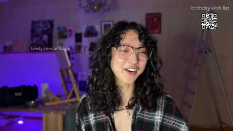 Media: Video of a young woman with curly black hair, glasses, and a plaid shirt, smiling. Background shows a dimly lit room with a painting easel, framed pictures, and purple lighting. Text overlay: \"birthday wish list\" and \"fanonly.com/u/luv.\