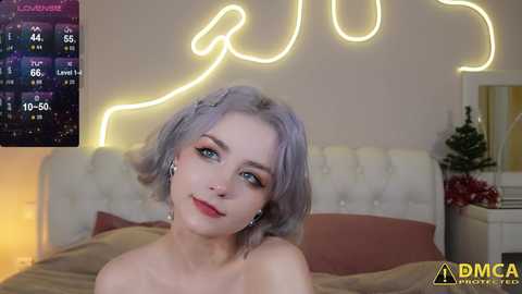 Media: A video of a fair-skinned woman with short, pastel purple hair, wearing a red lipstick smile, in a cozy bedroom with a neon \"LOVE\" sign and a digital clock.