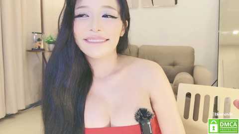 Media: Video of a smiling, light-skinned woman with long black hair, wearing a strapless red dress, holding a microphone, in a modern living room with beige furniture.