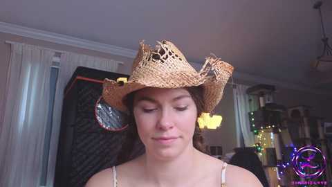 Media: Video of a young woman with fair skin and brown hair, wearing a straw cowboy hat with a decorative feather and a thin strap top. Background shows a beige room with curtains, a dark quilt, and a cat tower.