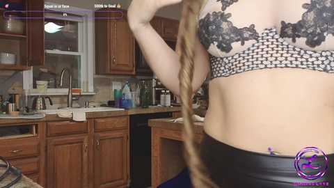 Media: Video of a light-skinned woman in a black lace bra, holding a long braid, in a kitchen with wooden cabinets and cluttered countertops.