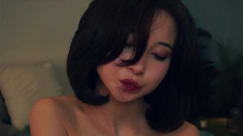 Media: A video of a young woman with short, dark hair and fair skin, looking pensive, wearing red lipstick, and possibly topless, in a dimly lit, blurred indoor setting.