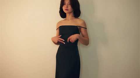 Media: Video of a slender, light-skinned Asian woman with shoulder-length black hair, wearing a strapless black dress, standing against a plain beige wall.