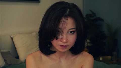 Media: Video of a young Asian woman with shoulder-length dark hair, light skin, and a slender build, topless, standing in a dimly lit room with beige walls and a bed with white pillows in the background.