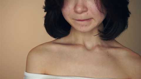 Media: A close-up video of a young woman with fair skin, shoulder-length black hair, and a white off-shoulder top. The background is a plain beige wall.