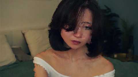 Media: Video of a young woman with shoulder-length, dark hair, fair skin, and green eyes, wearing a white off-shoulder top, sitting on a bed with beige pillows, dimly lit room background.