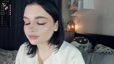 Media: Video of a young woman with fair skin, straight black hair, and closed eyes, wearing a white sweater, in a cozy bedroom with a lit fireplace, a bed with patterned sheets, and a dark curtain.