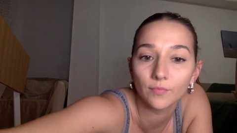 Media: A video of a young woman with light skin, brown eyes, and brown hair pulled back. She wears a gray tank top and silver hoop earrings. The background shows a plain room with a green couch and a wooden table.