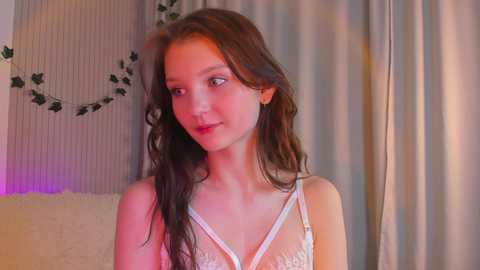 Media: Video of a young woman with long brown hair, fair skin, and a slender physique, wearing a sheer white lace bralette. Background features a cream-colored sofa, a floral wall hanging, and a hint of purple lighting.