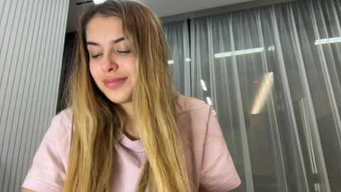 Media: Video of a young woman with long, blonde hair, wearing a light pink shirt, standing in a modern, well-lit room with gray curtains and vertical white paneling.