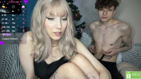 Media: Video of a pale, blonde woman with a thin body and medium-sized breasts, wearing black lingerie. A shirtless, curly-haired, slender man sits next to her. Background features a festive Christmas tree and text overlay.