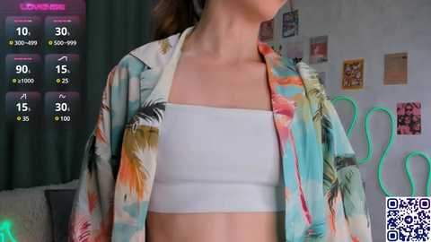 Media: Video of a young woman with light skin, wearing a white crop top and floral kimono, standing in a vibrant, artsy room with colorful wall art and a green curtain.