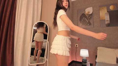 Media: Video of a slender, fair-skinned woman with long brown hair in a white crop top and frilled skirt, standing in a bedroom with a mirror, wall art, and a lamp.