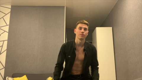 Media: A video of a shirtless, light-skinned man with short brown hair, wearing an unbuttoned black shirt, standing in a modern bedroom with grey walls and geometric-patterned wallpaper.