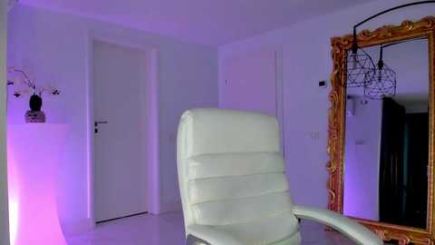 Media: Video of a minimalist, modern room with a white leather recliner chair, a white door, and a large mirror with ornate golden frame, lit by purple ambient lighting.