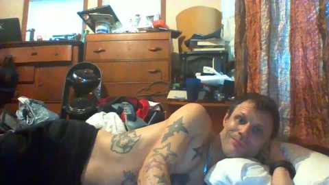 Media: Video of a shirtless, tattooed man with short dark hair, reclining on a bed in a cluttered bedroom with wooden furniture, a helmet, and patterned curtains.