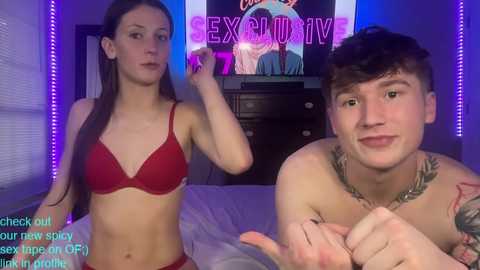 Video of a slender, fair-skinned woman in red lingerie and a young, shirtless, tattooed man, both smiling, in a dimly-lit bedroom.