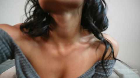 Media: Video of a woman with long, wavy black hair, wearing a loose grey off-shoulder sweater, exposing her collarbone. Background is a plain, light-colored wall.