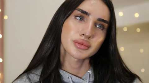 Video of a young woman with long, straight, dark brown hair, fair skin, and arched eyebrows, wearing a grey shirt. Background features blurred, warm-toned bokeh lights and a neutral wall.