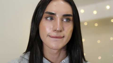 Media: Video of a young woman with straight, dark brown hair, fair skin, and light blue eyes. She has well-defined eyebrows, a slight smile, and wears a grey hoodie. Background features blurred bokeh lights.