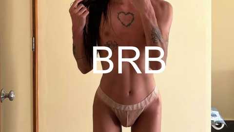 Video of a slender, topless woman with long dark hair, wearing beige panties, showcasing heart tattoos on her chest and arms. She stands in a beige room with a door and a white object in the background.