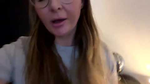 Media: A close-up video of a woman with long brown hair and glasses, wearing a white shirt, mouth open mid-sentence, blurred background.