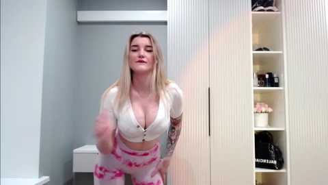 Media: A video of a blonde woman in a white crop top and pink tie-dye pants, standing in a modern, minimalist bathroom with white cabinets and a closed toilet.