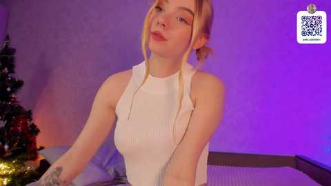 Media: A video of a young, fair-skinned, blonde woman with a slim physique wearing a white ribbed tank top, sitting in a room with purple lighting and a decorated Christmas tree.
