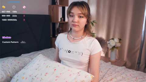 Media: Video of a young Asian woman with light skin, shoulder-length brown hair, and a white T-shirt with a floral pattern. She's sitting on a bed with floral bedding, in a cozy, softly lit room.