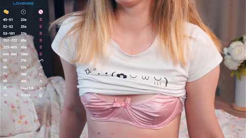 Media: Video of a fair-skinned woman with long blonde hair, wearing a white crop top tied at the waist, exposing a pink lace bra, standing indoors in a bedroom with a floral bedspread.