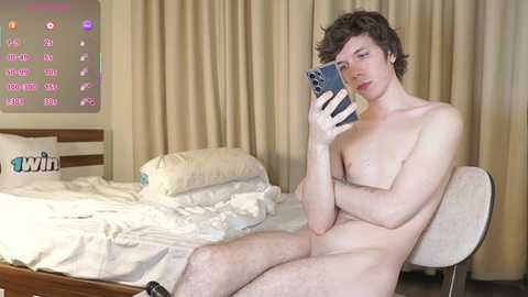 Media: Video of a young, slender, fair-skinned man with curly brown hair, nude, taking a selfie in a hotel room with a messy bed, beige curtains, and a TV displaying a \"win\" logo.