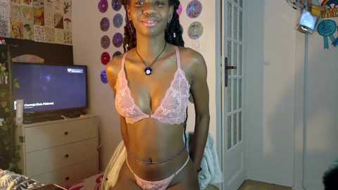 Media: Video of a young Black woman with dark skin and long braided hair, wearing pink lace lingerie. She stands in a cluttered bedroom with a TV, art posters, and a bed visible.