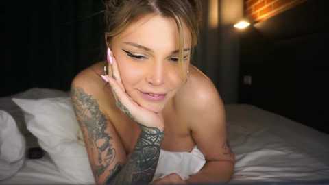 Media: Video of a topless, tattooed woman with blonde hair, lying on a bed in a dimly lit room, smiling.