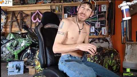 Media: Video of a shirtless, tattooed man with a beard and jeans, sitting on a gaming chair, surrounded by gaming and motorcycle accessories in a cluttered room.