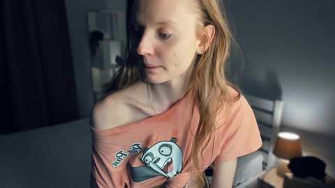 Media: Video of a young woman with fair skin, long auburn hair, wearing a peach off-shoulder T-shirt with cartoon character \"Adventure Time.\" She sits in a dimly lit room with a bed, chair, and lamp in the background.