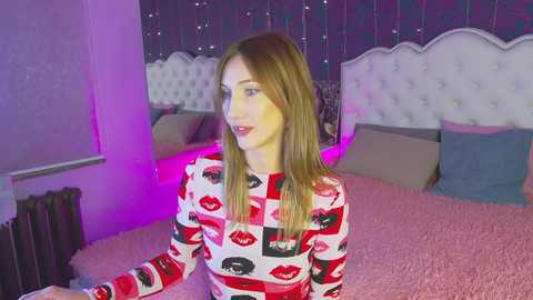 Media: Video of a young, fair-skinned woman with long blonde hair, wearing a red lipstick print top, sitting on a pink bed with a tufted white headboard. Background features a purple light, floral wallpaper, and a radiator.