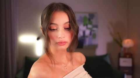 Media: Video of a young woman with fair skin and brown hair, wearing a white off-shoulder top, standing indoors in a softly lit room with a green bed and abstract art on the wall.
