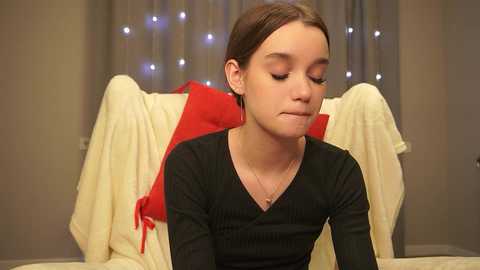 Media: Video of a young girl with fair skin, brown hair, and a black V-neck top, sitting contemplatively on a beige sofa with a red towel and white blanket behind her.