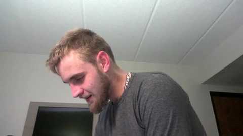 Media: Video of a young man with short, light brown hair and a beard, wearing a gray t-shirt, indoors with white ceiling and dark wooden door.