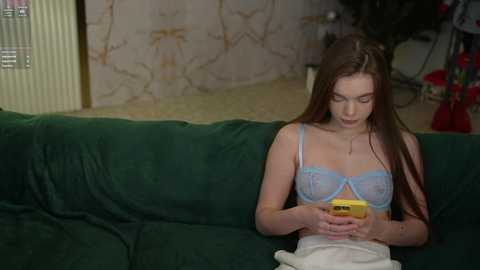 Media: Video of a young Caucasian woman with long brown hair, wearing a blue lace bra, sitting on a green couch, texting on a yellow phone, in a dimly lit room with a floral wallpaper.