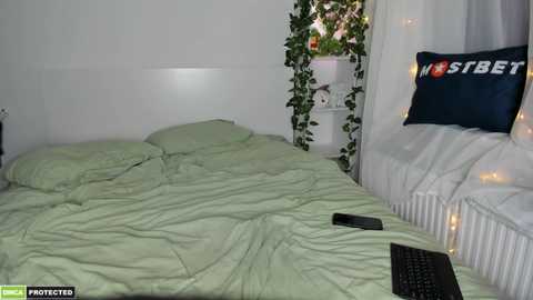 Media: A video of a cozy, white-walled bedroom with a green bedspread, a dark blue pillow with \"SBRY\" text, fairy lights, and a black laptop on the bed.